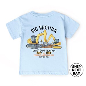 A light blue t-shirt with a graphic design depicting construction equipment and the text "BIG BROTHER UNDER CONSTRUCTION ESTIMATED COMPOSITION DATE December 2015".