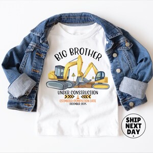 a white t-shirt with a graphic design depicting construction equipment and the text "BIG BROTHER UNDER CONSTRUCTION ESTIMATED COMPLETION DATE DECEMBER 2025". The t-shirt is placed on top of a denim jacket, and there is a shipping label next to it.