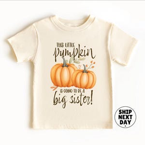 以下が含まれることがあります： 「This little pumpkin is going to be a big sister!」の文字と、オレンジ色のカボチャ2つの水彩イラストが描かれた、クリーム色の幼児用Tシャツ。半袖で丸首です。