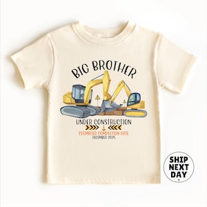 Puede incluir: Camiseta crema para niños pequeños con un gráfico de temática de construcción. El diseño presenta excavadoras amarillas, el texto "BIG BROTHER UNDER CONSTRUCTION" y "ESTIMATED COMPLETION DATE DECEMBER 2025".
