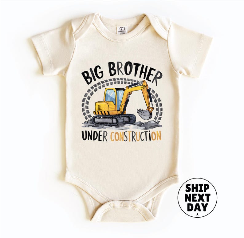 A baby onesie with a graphic of a yellow excavator and the text "BIG BROTHER UNDER CONSTRUCTION" printed on it.
