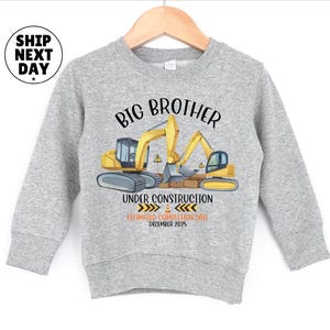 A gray sweatshirt with a graphic design featuring a construction site with a crane and a message that reads "BIG BROTHER UNDER CONSTRUCTION ESTIMATED COMPLETION DATE DECEMBER 2015".