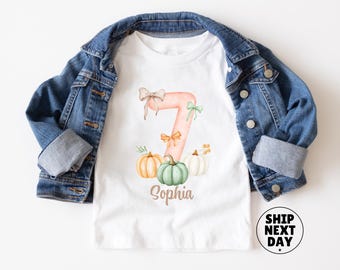 Könnte beinhalten: Ein weißes T-Shirt mit einer großen pfirsichfarbenen Zahl sieben, verziert mit kleinen Schleifen und drei Kürbissen in Orange, Grün und Weiß. Der Name Sophia steht unter der Zahl. Über dem T-Shirt liegt eine blaue Jeansjacke.