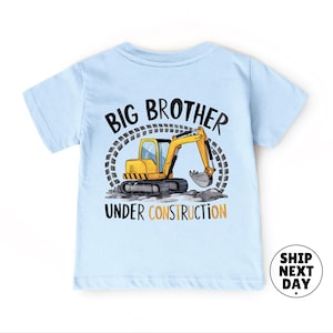 A light blue t-shirt with a graphic design depicting a yellow construction vehicle and the text "BIG BROTHER UNDER CONSTRUCTION" printed on it.