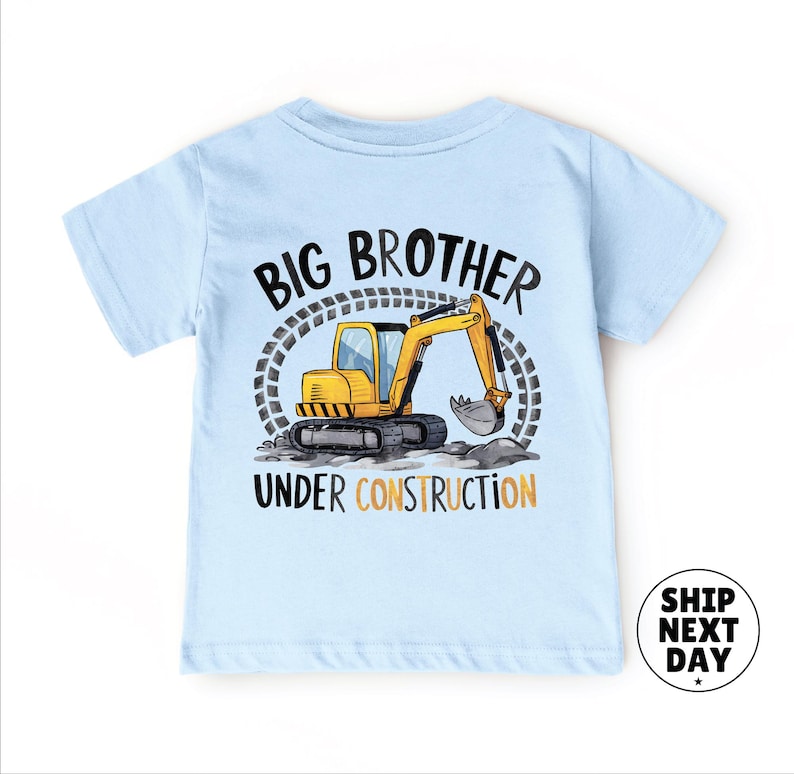 A light blue t-shirt with a graphic design depicting a yellow construction vehicle and the text "BIG BROTHER UNDER CONSTRUCTION" printed on it.