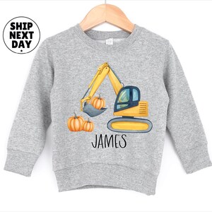Pode incluir: Camisola cinza claro com um design de escavadeira amarela, abóboras e o nome "JAMES" em preto. A camisola tem mangas compridas e gola redonda.