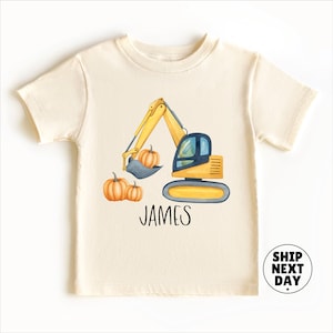 May include: Cream-colored short-sleeve t-shirt featuring a watercolor illustration of a yellow excavator with a blue cab and pumpkins. The name "JAMES" is printed below the excavator. A circular "SHIP NEXT DAY" graphic is in the lower right.