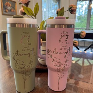 May include: Two insulated tumblers, one sage green and one light pink, with white lids and handles. Each tumbler features the text "The joy of the Lord is my strength" with a floral design.