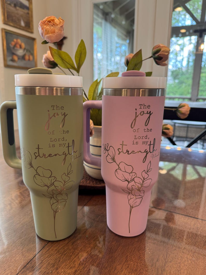 May include: Two insulated tumblers, one sage green and one light pink, with white lids and handles. Each tumbler features the text "The joy of the Lord is my strength" with a floral design.