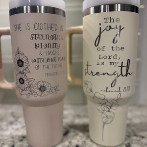 May include: Two insulated tumblers with lids. One is light pink with a sunflower design and the text "She is clothed in strength & dignity & laughs without fear of the future." The other is cream-colored with the text "The joy of the Lord is my strength."
