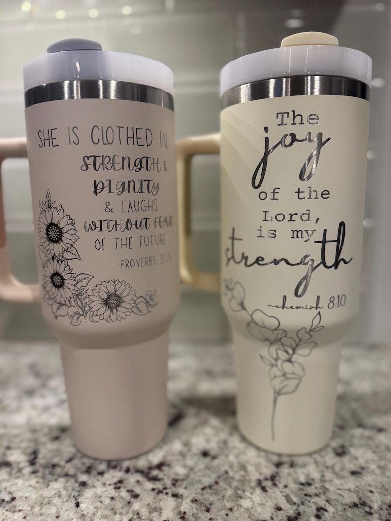 May include: Two insulated tumblers with lids. One is light pink with a sunflower design and the text "She is clothed in strength & dignity & laughs without fear of the future." The other is cream-colored with the text "The joy of the Lord is my strength."