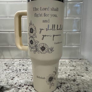 May include: A beige insulated tumbler with a handle, a silver rim, and a white lid. The tumbler features the text "The Lord shall fight for you, and ye shall hold your peace" with floral accents. The name "Rebekah" is also printed on the tumbler.