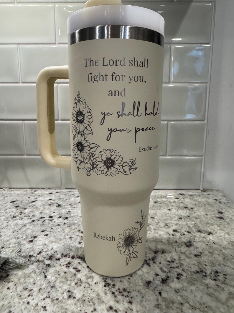 May include: A beige insulated tumbler with a handle, a silver rim, and a white lid. The tumbler features the text "The Lord shall fight for you, and ye shall hold your peace" with floral accents. The name "Rebekah" is also printed on the tumbler.