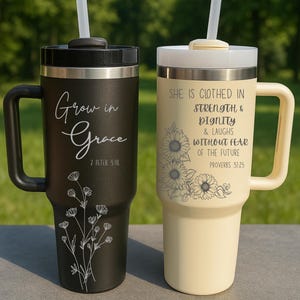 May include: Two insulated travel mugs with handles and straws. One is black with the words "Grow in Grace" and a floral design. The other is cream-colored with the text "She is clothed in strength & dignity" and a sunflower design.