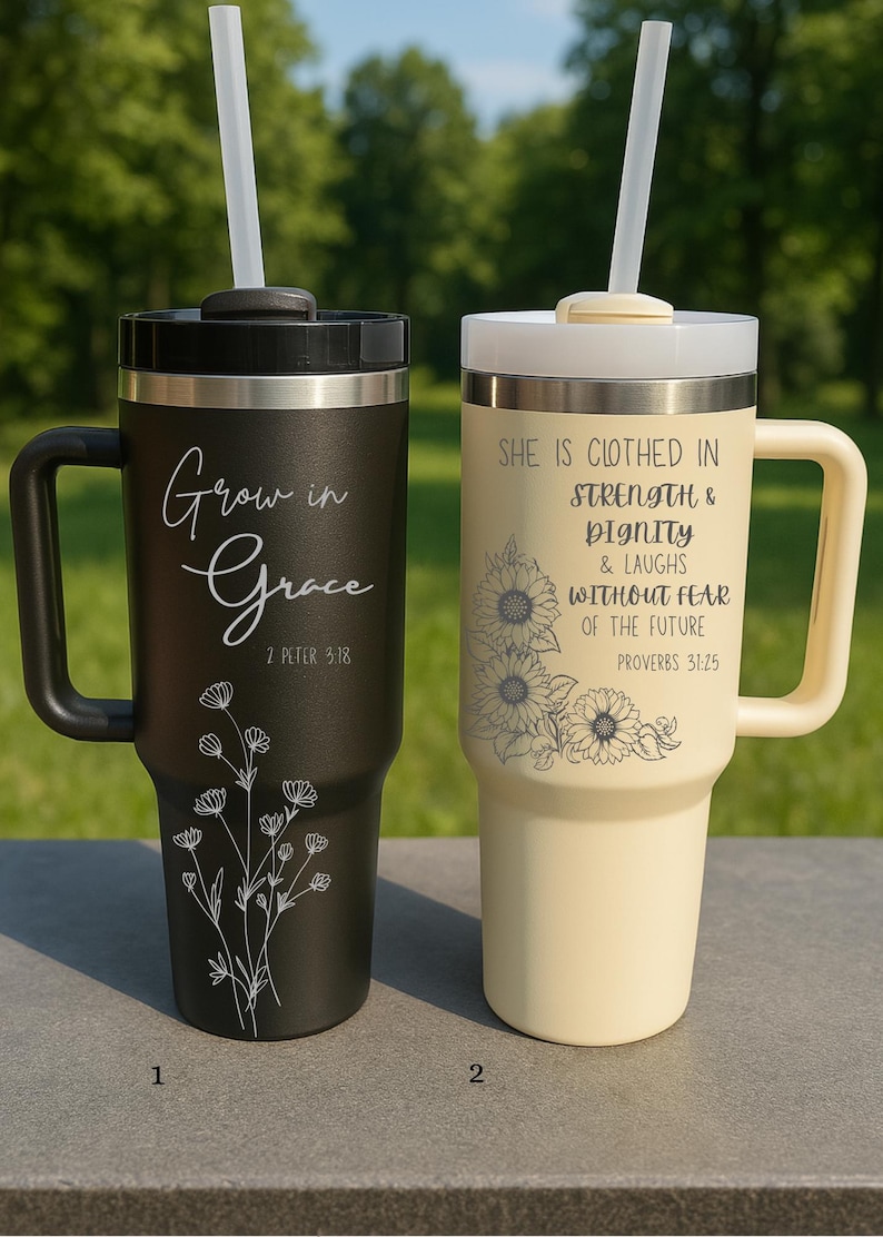 May include: Two insulated travel mugs with handles and straws. One is black with the words "Grow in Grace" and a floral design. The other is cream-colored with the text "She is clothed in strength & dignity" and a sunflower design.