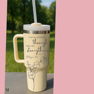 May include: A cream-colored tumbler with a handle and a white lid, featuring a straw. The tumbler has the text "I can do all things through him who strengthens me. Philippians 4:13" and a floral design.