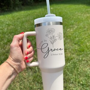 May include: A cream-colored tumbler with a white lid and handle, featuring the text "Grow in Grace" and a floral design. Includes a straw. The tumbler is held against a backdrop of green grass.