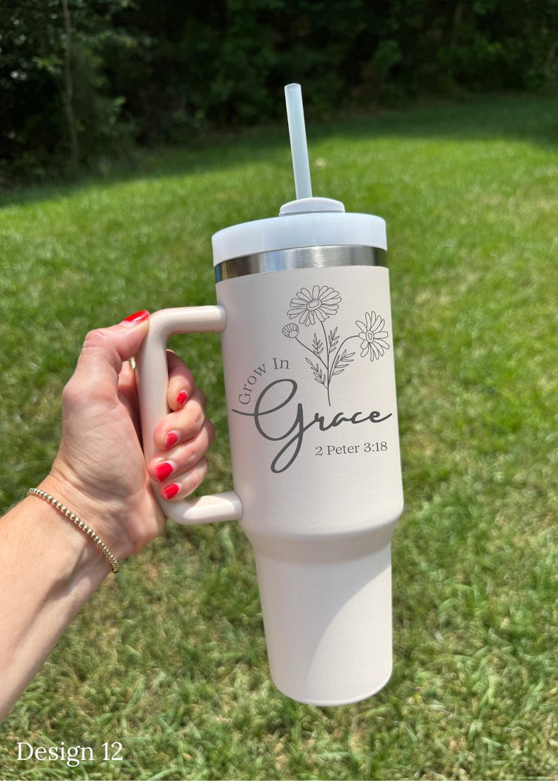 May include: A cream-colored tumbler with a white lid and handle, featuring the text "Grow in Grace" and a floral design. Includes a straw. The tumbler is held against a backdrop of green grass.