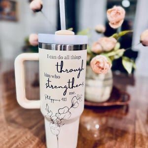 May include: Cream-colored insulated tumbler with a handle, lid, and straw. The tumbler features the text "I can do all things through him who strengthens me. Philippians 4:13" and a floral design. The handle and straw are light beige.
