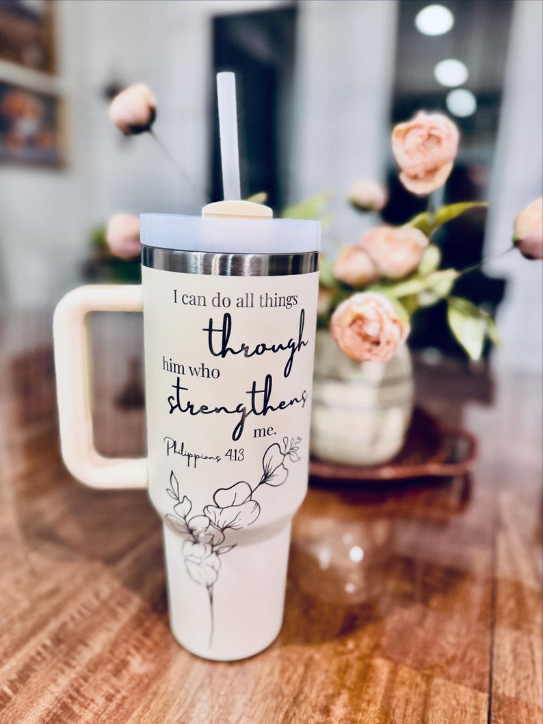 May include: Cream-colored insulated tumbler with a handle, lid, and straw. The tumbler features the text "I can do all things through him who strengthens me. Philippians 4:13" and a floral design. The handle and lid are a matching cream color.