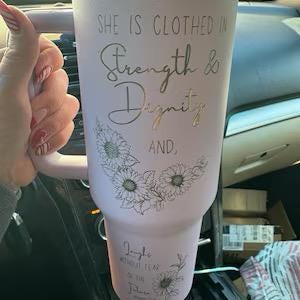 May include: A light pink tumbler with a handle, featuring the text "SHE IS CLOTHED IN Strength & Dignity AND..." and sunflower illustrations. The tumbler is designed to hold beverages.