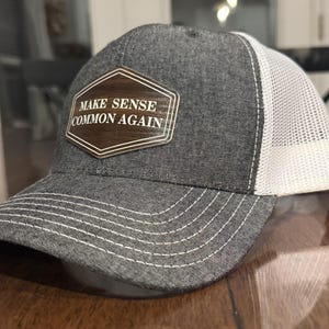 May include: A gray trucker hat with a white mesh back and a curved brim. The front of the hat features a wooden patch with the words "MAKE SENSE COMMON AGAIN" in white lettering.