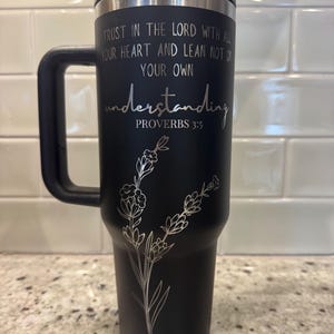 May include: Black insulated travel mug with a black handle and lid. The mug features the text "TRUST IN THE LORD WITH ALL YOUR HEART AND LEAN NOT ON YOUR OWN understanding PROVERBS 3:5" and a floral design.
