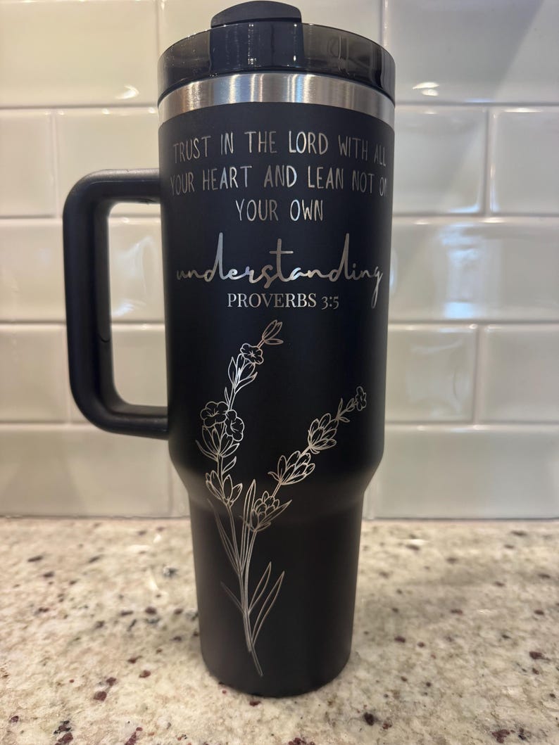 May include: Black insulated travel mug with a black handle and lid. The mug features the text "TRUST IN THE LORD WITH ALL YOUR HEART AND LEAN NOT ON YOUR OWN understanding PROVERBS 3:5" and a floral design.