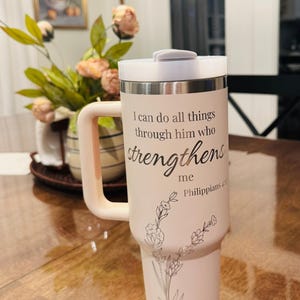 May include: A light pink travel mug with a white lid and handle. The mug features the text "I can do all things through him who strengthens me Philippians 4:13" and a floral design. The mug is designed to hold approximately 30 ounces.