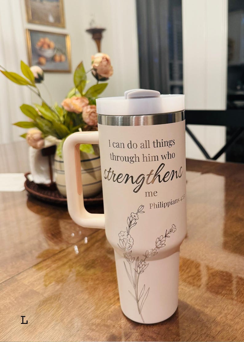 May include: A light pink travel mug with a white lid and handle. The mug features the text "I can do all things through him who strengthens me Philippians 4:13" and a floral design. The mug is designed to hold approximately 30 ounces.