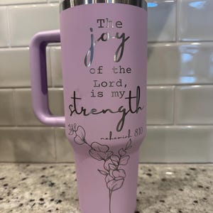 May include: A lavender insulated tumbler with a white and silver lid and handle. The text "The joy of the Lord is my strength" is displayed in silver script, along with a floral design and the reference "nehemiah 8:10".