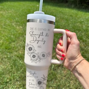 May include: A light beige tumbler with a white lid and handle, featuring the text "She is clothed in Strength & Dignity" and "Laughs without fear of the future". The design includes sunflower illustrations and a straw.