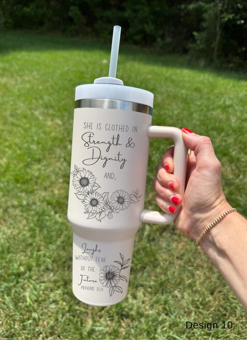 May include: A light beige tumbler with a white lid and handle, featuring the text "She is clothed in Strength & Dignity" and "Laughs without fear of the future". The design includes sunflower illustrations and a straw.