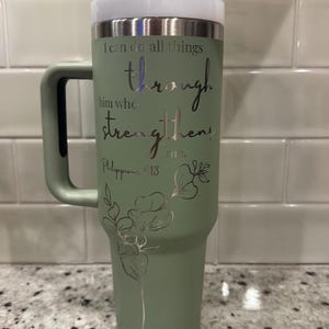 May include: A sage green tumbler with a handle, a white lid, and a clear straw. The tumbler has the text "I can do all things through him who strengthens me. Philippians 4:13" in silver script, along with a floral design.