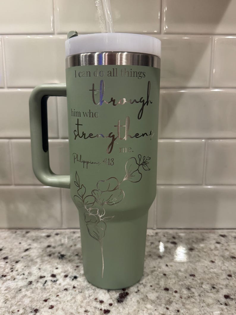 May include: A sage green tumbler with a handle, a white lid, and a clear straw. The tumbler has the text "I can do all things through him who strengthens me. Philippians 4:13" in silver script, along with a floral design.