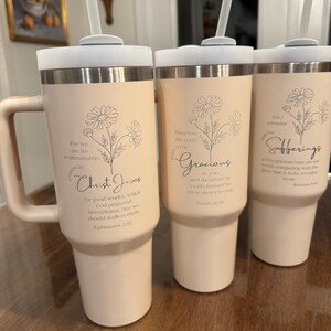 May include: Three insulated tumblers with lids and straws. The tumblers are a light peach color with a silver band at the top and bottom. Each tumbler has a floral design and a religious quote in black script. The tumblers have a handle.