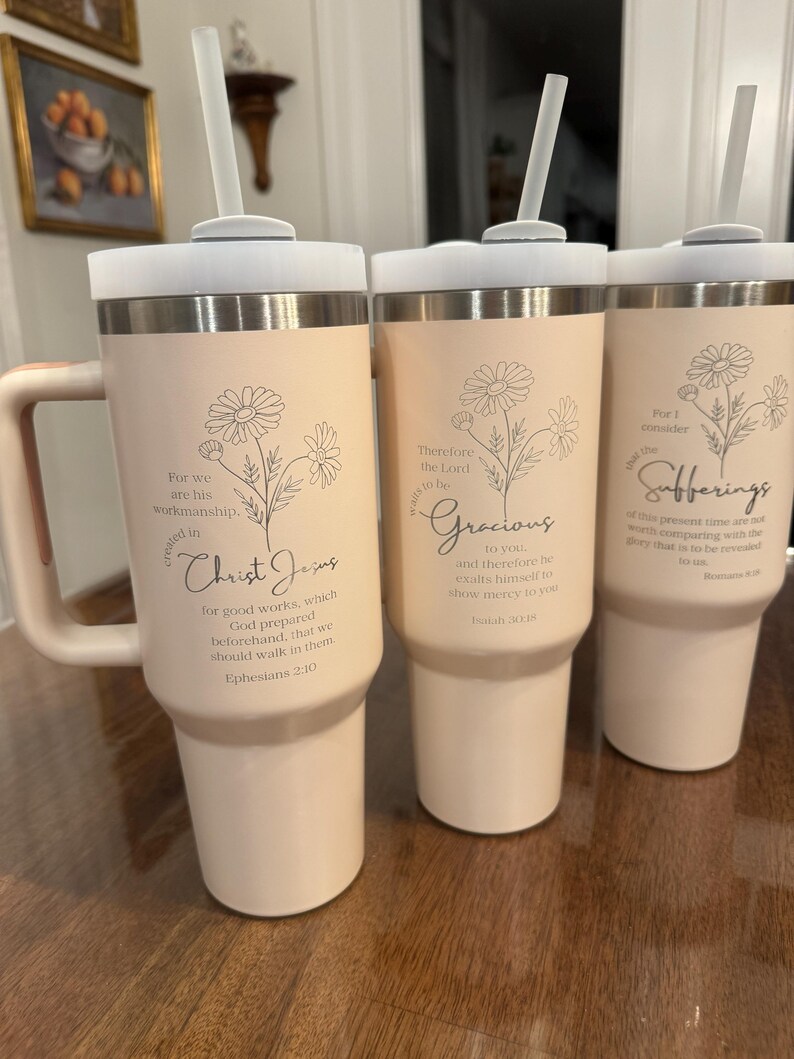 May include: Three insulated tumblers with lids and straws. The tumblers are a light peach color with a silver band at the top and bottom. Each tumbler has a floral design and a religious quote in black script. The tumblers have a handle.