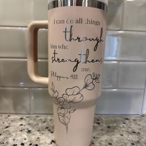 May include: A light pink insulated tumbler with a handle and a lid. The tumbler features the text "I can do all things through him who strengthens me. Philippians 4:13" in silver lettering, along with a floral design.