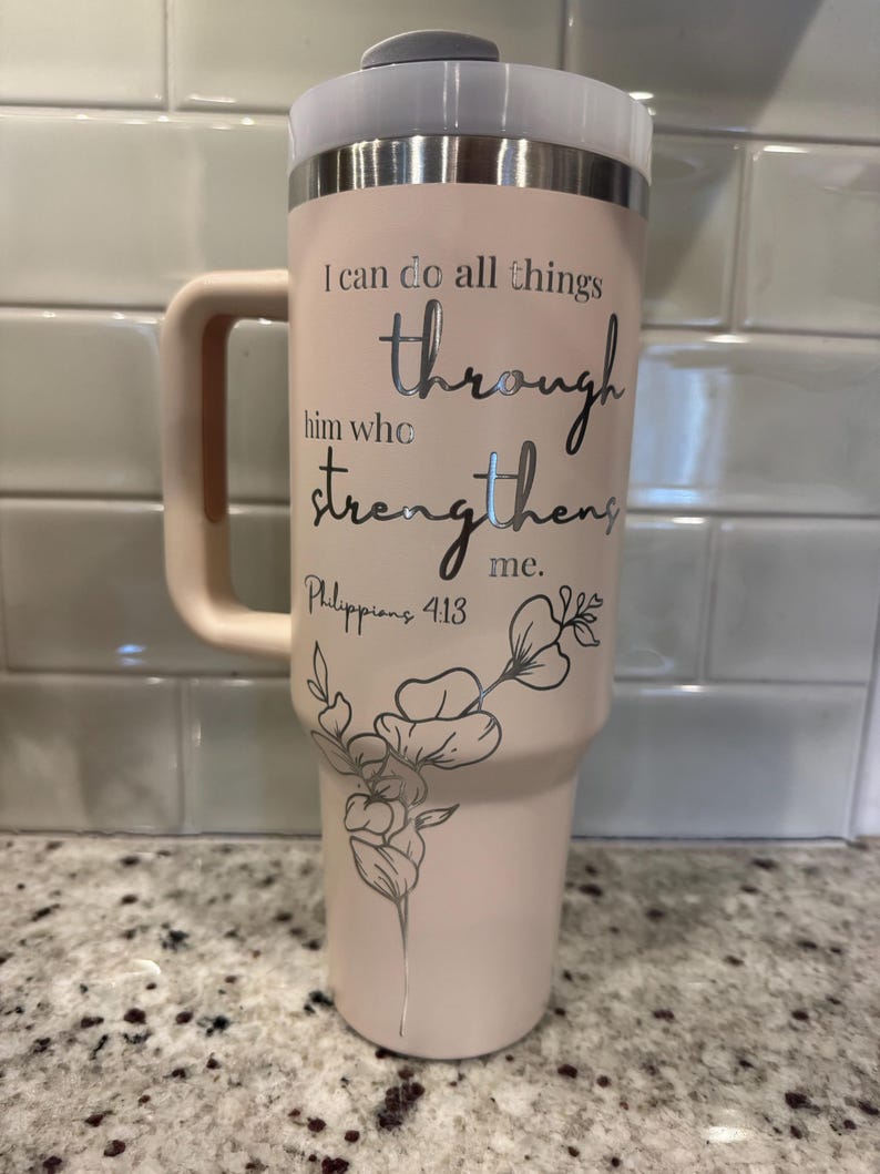 May include: A light pink insulated tumbler with a handle and a lid. The tumbler features the text "I can do all things through him who strengthens me. Philippians 4:13" in silver lettering, along with a floral design.