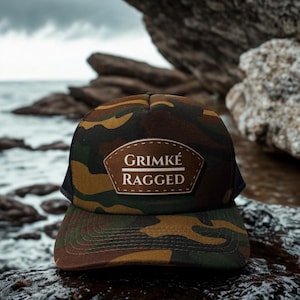 May include: Camouflage-patterned trucker hat with a brown leather patch that reads "Grimké Ragged." The hat features a curved brim and a black mesh back. The hat is sitting on a wet, rocky surface.