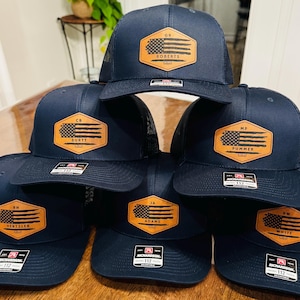 May include: A stack of navy blue trucker hats with a mesh back. Each hat features a brown leather patch with a distressed American flag design and a name. The hats are adjustable and have a Richardson 112 tag.