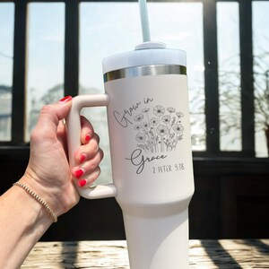 May include: A white insulated tumbler with a handle, lid, and straw. The tumbler features the text "Grow in Grace" and a floral design. The background includes a window and sunlight.