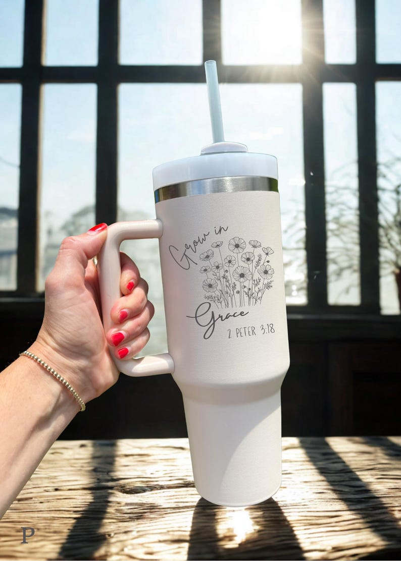 May include: A white insulated tumbler with a handle, lid, and straw. The tumbler features the text "Grow in Grace" and a floral design. The background includes a window and sunlight.