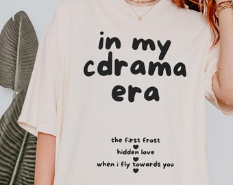 May include: Cream-colored t-shirt with the text "in my cdrama era" in black, along with the phrases "the first frost", "hidden love", and "when i fly towards you". The shirt has short sleeves and a relaxed fit.