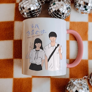 以下が含まれることがあります： ピンクの持ち手が付いた白いセラミックマグ。2人のイラストが描かれています。マグには英語と別の言語でテキストが書かれています。背景にはディスコボールとチェック柄があります。