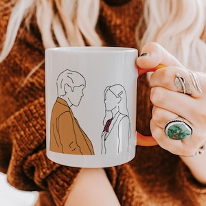 Peut inclure: Mug en céramique blanche avec une illustration minimaliste au trait de deux personnages se faisant face. L'intérieur et la anse de la tasse sont rouges. L'illustration représente un homme en manteau marron et une femme en blazer blanc.