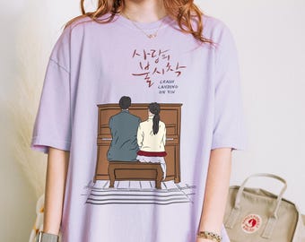 May include: A light purple t-shirt featuring a graphic of a couple at a piano. The text above the graphic is in Korean, with the English text "CRASH LANDING ON YOU" below. The shirt is a soft, comfortable material.