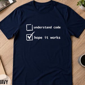 Può includere: T-shirt blu navy con testo bianco che recita "understand code" e "hope it works" con un segno di spunta accanto alla seconda frase. La maglietta è esposta su una superficie di legno.