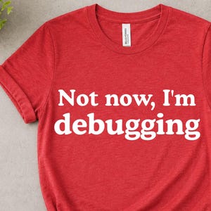 May include: A heather red t-shirt with the text "Not now, I'm debugging" in white. The shirt is knotted at the bottom and has rolled-up sleeves. A small plant, a camera, and a cup of coffee are in the background.