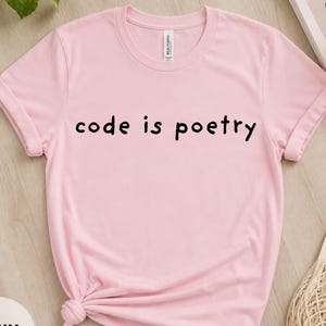 Könnte beinhalten: Ein hellrosa T-Shirt mit dem schwarzen Aufdruck "code is poetry". Das Shirt ist unten geknotet und hat hochgekrempelte Ärmel. Ein kleines Etikett ist am Halsausschnitt sichtbar. Das Wort "PINK" ist in einem Kreis unten aufgedruckt.